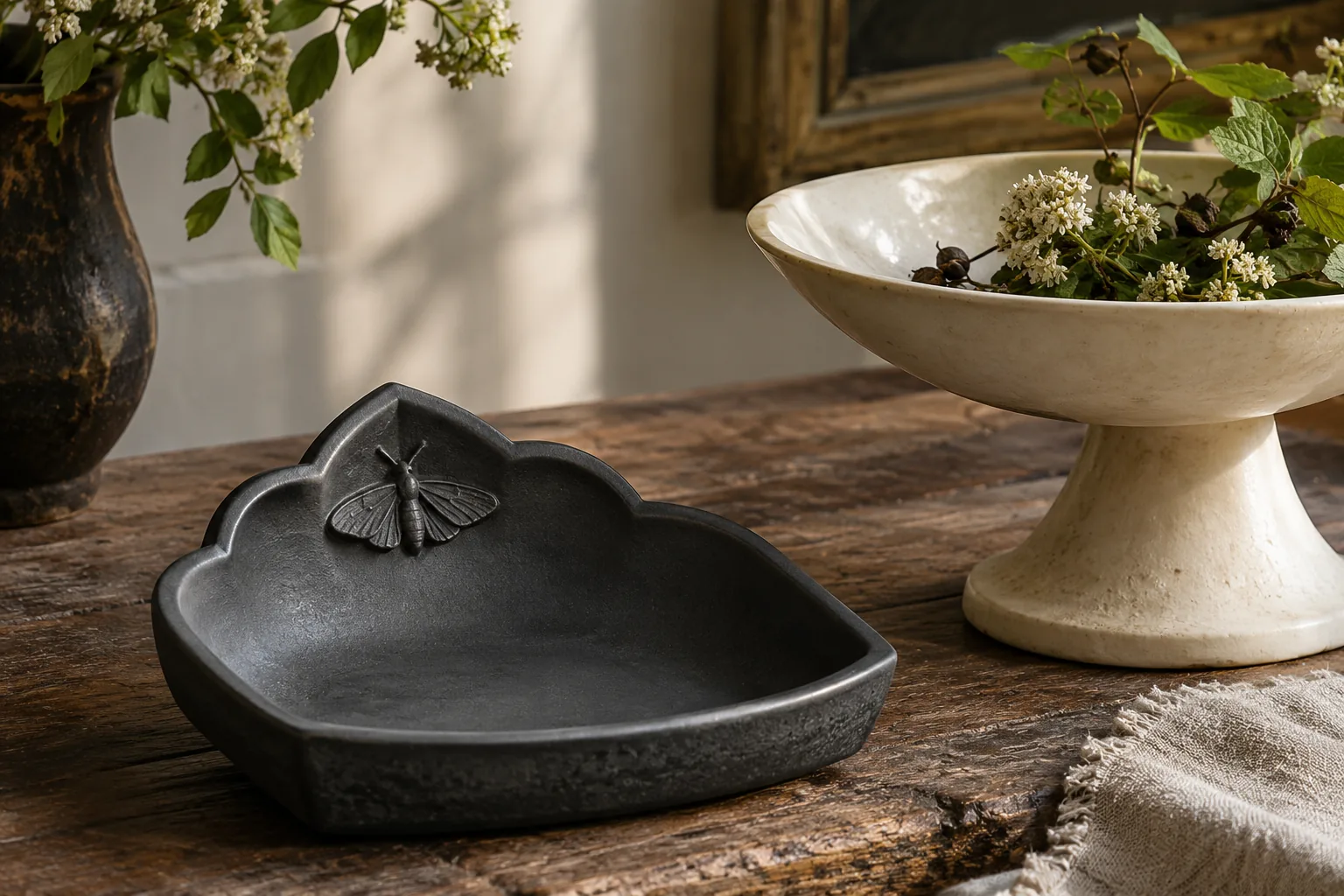 A matte charcoal ceramic dish with a broad gothic arch edge and a tiny sculptural moth relief beside an ivory pedestal bowl, moody natural daylight
